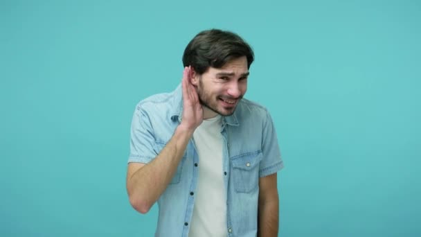 病気や聴覚の問題 ジーンズのシャツの中で注目すべき髭の男が耳の近くに手を取り合って ゴシップ 秘密のニュースを注意深く聞いています 青い背景に隔離された屋内スタジオで — ストック動画
