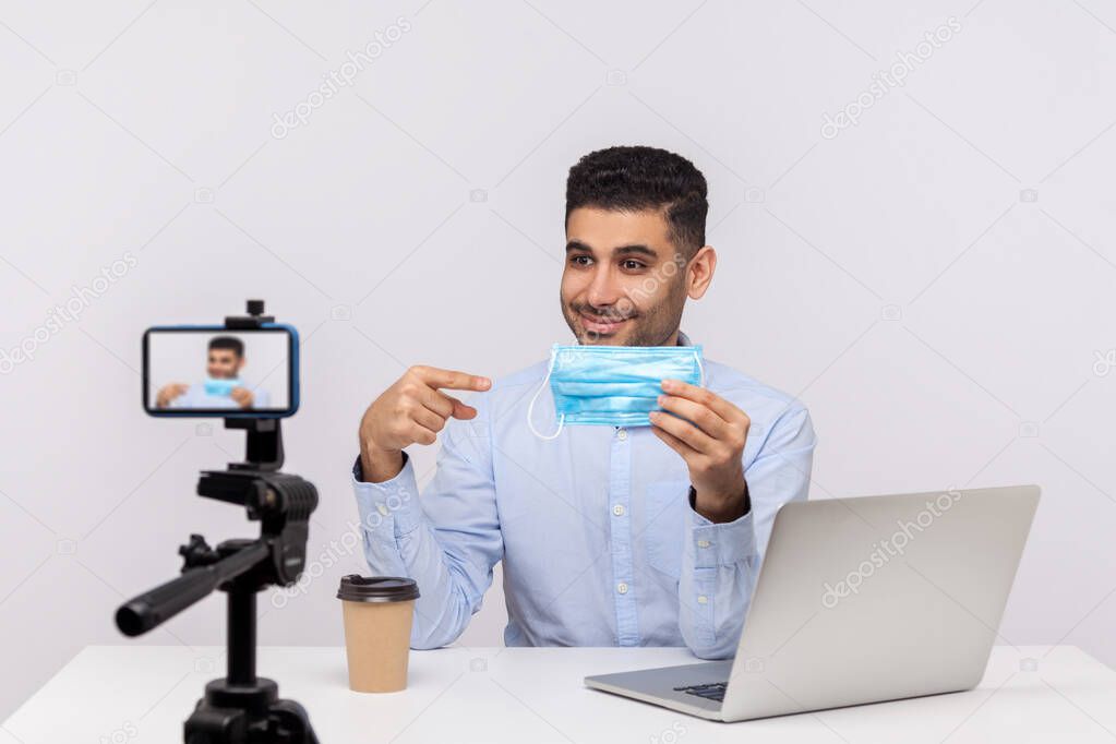 Positive man pointing at medical mask and broadcasting about coronavirus, making video giving tips on using protect filter against contagious disease, 2019-ncov, flu outbreak. indoor studio shot