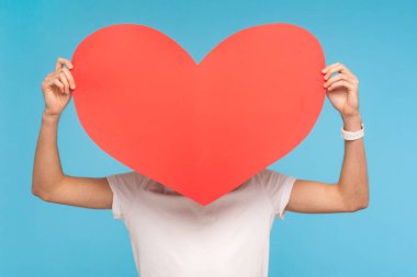 Sevgililer Günü için isimsiz tebrik! Beyaz tişörtlü adam büyük kırmızı kalbin arkasında duruyor, yüzünü gizliyor ve romantik bir hediye tutuyor, aşkını ilan ediyor. Stüdyo çekimi mavi arkaplanda izole edildi