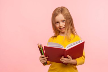 Eğitim, ilkokul. Kameraya gülümseyen sevimli küçük bir kızın portresi, elinde büyük bir kitap ve kalemler, yaratıcı çocuk yeteneklerinin gelişimi. Pembe arka planda kapalı stüdyo çekimleri