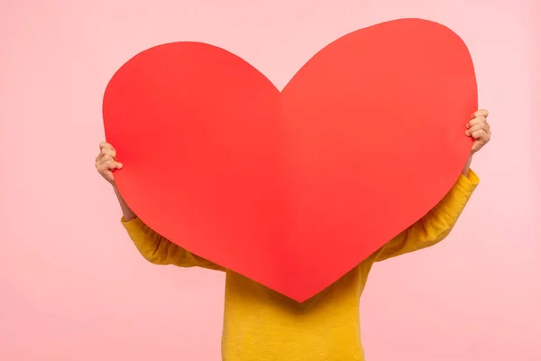 大きな赤い紙の心の後ろに隠れて認識できない人は 愛の愛情の愛情の象徴を保持 匿名の慈善とケアの概念は 心臓の健康 ピンクの背景に隔離された屋内スタジオで — ストック写真