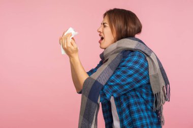 Mevsimsel grip. Eşarba sarılı hasta gömlekli, burnu akan, peçeteye hapşıran, viral enfeksiyon belirtileri gösteren hasta kızın yan görüntüsü. Pembe arka planda kapalı stüdyo çekimleri