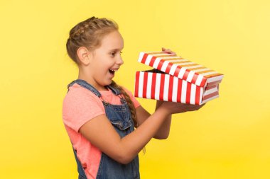Portrait of glad curious little girl with braid in denim overalls peeking into gift box and laughing from happiness, enjoying best surprise, holiday bonus. studio shot isolated on yellow background clipart