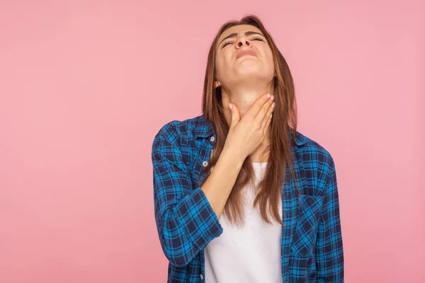 Throat pain. Portrait of flu sick girl in checkered shirt touching her inflamed throat and grimacing from pain, suffering tonsillitis, thyroid disorders. indoor studio shot isolated on pink background