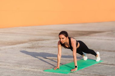 Dar spor giysili, siyah pantolonlu, üstsüz güzel esmer kadın yoga yapıyor, Phalakasana 'da yüksek tahta duruşunda duruyor, kaslarını ve gücünü geliştiriyor. Sağlık hizmetleri, dışarıda spor faaliyetleri