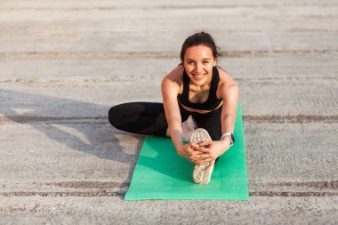 Güler yüzlü, spor kıyafetli, siyah pantolonlu ve üstsüz atletik bir kadın yoga yapıyor, dizinden dizine poz veriyor ayak parmaklarına dokunuyor, bacak ve sırt kaslarını esnetiyor. Sağlık hizmetleri, dışarıda spor faaliyetleri