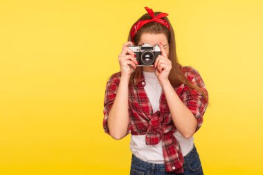 Kareli gömlekli ve bantlı bir kızın portresi eski moda bir kamerayla fotoğraf çekiyor, gezgin fotoğraf çekerek geziyor. Retro 50 'lerin tarzı, stüdyo çekimi sarı arka planda izole edilmiş.