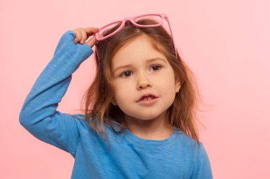 Muhteşem güzel, tapılası küçük bir kızın portresi. Pembe gözlüklerini çıkarıp rüya gibi bir ifadeyle, şık bir çocukla, moda konseptiyle bakıyor. Pembe arka planda kapalı stüdyo çekimleri