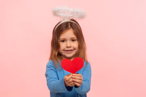 Portrait of adorable cute preschool girl with angelic nimbus holding paper heart and smiling to camera, little cupid for Valentines day sharing love. indoor studio shot isolated on pink background