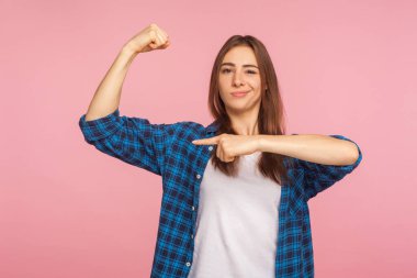 Bak, ben güçlüyüm! Kareli gömlekli, kibirli ve başarılı bir kızın portresi pazıları gösteriyor ve kibirli bir ifadeyle, güçlü ve kendine güvenle bakıyor. Stüdyo çekimi pembe arkaplanda izole edildi