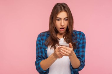 Mobil bağlantı. Sosyal ağda kareli tişört okuyup cep telefonuyla sohbet eden şaşırmış kızın portresi. Pembe arka planda kapalı stüdyo çekimleri