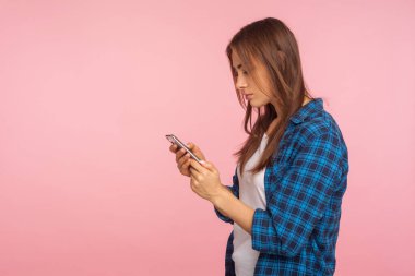 Mobil bağlantı. Sosyal ağda cep telefonu kullanarak, sohbet ederek, mesaj yazarak, tişört giyen ciddi bir kızın yan görüntüsü. Pembe arka planda izole edilmiş kapalı stüdyo çekimi, kopyalama alanı