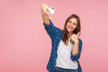 Mükemmel mobil iletişim. Kareli tişörtlü neşeli bir kızın portresi gülümseyen ve başparmağını kaldıran bir jest gibi video konuşmaları, online sohbetler. Stüdyo çekimi, izole