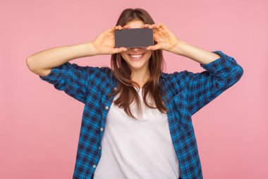 Mobil cihazın reklamı. Kareli tişörtlü mutlu kız gözlerini akıllı telefonla kapatıyor, yüzünü yarım yamalak saklıyor ve kameraya gülümsüyor, neşe saçıyor. Pembe arka planda kapalı stüdyo çekimleri