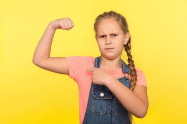 Bak, ben güçlüyüm! Saç örgülü, bisepsi olan cesur küçük bir kızın portresi. Sağlıklı olmak için güç ve kendine güven gösteren bir çocuk. Sarı arka planda izole edilmiş kapalı stüdyo çekimi