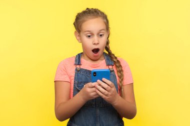 Şok olmuş küçük bir kızın portresi cep telefonundan şaşırtıcı mesajlar okuyor, sosyal ağda sohbet ediyor, çocuklar için iyi bir cep telefonu uygulaması karşısında hayrete düşüyor. Sarı arka planda izole edilmiş kapalı stüdyo çekimi