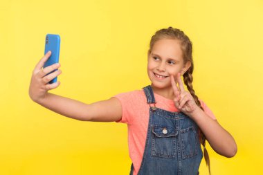 Kot pantolon giymiş sevimli küçük bir kızın portresi zafer ya da barışı simgelerken ebeveynlerle video görüşmesi yapıyor, cep telefonundan selfie çekiyor. Stüdyo çekimi sarı arkaplanda izole edildi