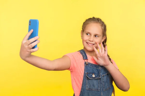 穿着粗斜纹棉布工作服的可爱小女孩在与父母进行视频通话 在手机上自拍时 穿着工作服给自己做手势 在黄色背景下隔离的室内拍摄 — 图库照片