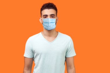 Tıbbi maskeli, sıradan beyaz tişörtlü, elleri aşağıda kameraya odaklanmış ve kendinden emin bir şekilde duran yakışıklı bir adamın portresi. Turuncu arka planda kapalı stüdyo çekimleri