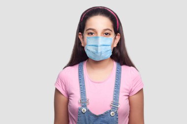 Tıbbi maskeli bir kız portresi, günlük tarz, pembe tişört ve mavi kot tulumlu dikilip bakıyor. Gri arka planda izole edilmiş kapalı stüdyo görüntüleri..