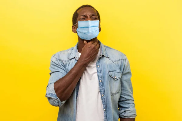 Portrait of upset man with medical mask holding his neck feeling unwell, suffering sore throat, inflammation and flu symptoms, medical concept. indoor studio shot isolated on yellow background