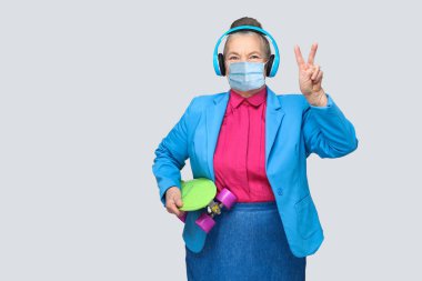Renkli tıbbi maskeli, yeşil kaykay taşıyan mavi kulaklıklı yaşlı bir kadın. Müzik dinliyor, barış işareti yapıyor ve kameraya bakıyor. Kapalı, gri arka planda izole.