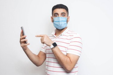 Beyaz tişörtlü, ayakta duran, elinde telefon tutan, işaret eden, şaşırmış yüzlü, iri gözlü genç bir adamın portresi. İçerde, izole edilmiş, stüdyoda çekilmiş, beyaz arka plan, kameraya bakıyor.