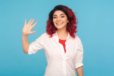 Merhaba, hoş geldiniz! Havalı kızıl saçlı mutlu hippi kadının portresi. Merhaba diyor ve kameraya gülümsüyor. Neşeyle konuksever ev sahibesi buluşması. Mavi arka planda kapalı stüdyo çekimleri