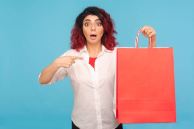 Süslü kızıl saçlı, büyük paketleri gösteren ve hafta sonu alışverişinin başarılı olmasına hoş bir şekilde şaşırmış görünen alışveriş düşkünü hippi kadının portresi. Stüdyo çekimi mavi arkaplanda izole edildi
