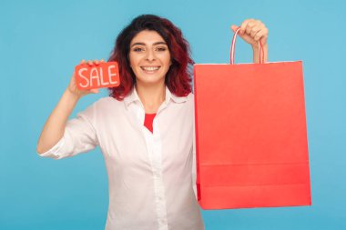 Moda mağazasında indirim. Süslü kızıl saçlı neşeli hippi kadın satılık yazılar ve alışveriş torbaları gösteriyor. Kara Cuma 'nın tadını çıkarıyor. Düşük fiyatlar. Mavi arka planda kapalı stüdyo çekimleri
