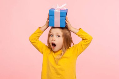 Portrait of astonished little girl holding gift box on head with expression of much surprise and amazement, shocked by birthday present, holiday bonus. indoor studio shot isolated on pink background clipart