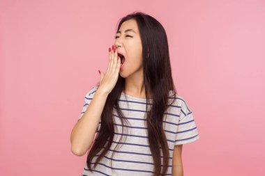 Uykulu, yorgun, esmer, çizgili tişörtlü, ağzı eliyle esneyen, uykulu, yorgun ve enerjisiz bir kız portresi. Pembe arka planda kapalı stüdyo çekimleri