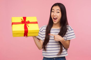 Awesome birthday present! Amazed satisfied pretty girl with brunette hair in striped t-shirt pointing at gift box and looking with surprised expression. indoor studio shot isolated on pink background clipart