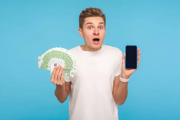 オンライン銀行 支払い ショックの驚き 金融取引アプリの広告を表現し ユーロ紙幣やスマートフォンを保持Tシャツの素晴らしい男 スタジオショットは青の背景に隔離され — ストック写真