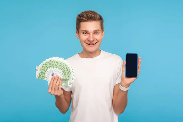 支払いと振込 オンライン銀行 ユーロ紙幣とスマートフォンを持っているTシャツの幸せな笑顔の男 金融取引アプリの広告 青い背景に隔離された屋内スタジオで — ストック写真