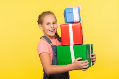Portrait of delighted little girl with braid in denim overalls holding lot of gift boxes and smiling to camera, enjoying many presents on her birthday. indoor studio shot isolated on yellow background clipart