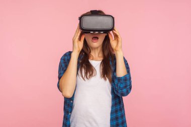 Damalı tişörtlü, VR kulaklık takmış, şok olmuş yüz ifadesiyle sanal gerçeklik oyunu oynayan, yenilikçi teknolojiye şaşırmış oyuncu kızın portresi. Kapalı stüdyo çekimi, izole.