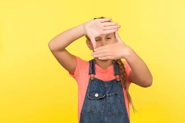 Portrait of little girl in denim overalls looking through photo frame hand gesture, focusing zooming picture, child observing world with interest. indoor studio shot isolated on yellow background