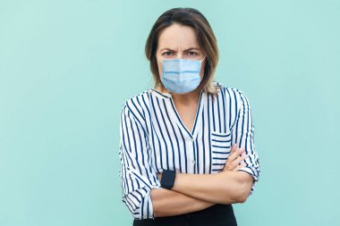 Cerrahi maske takmış, kolları çapraz duran ve kaşlarını çatmış kameraya bakan kızgın orta yaşlı bir kadının portresi. İlaç, sağlık hizmeti konsepti. Mavi arka planda kapalı stüdyo çekimleri.