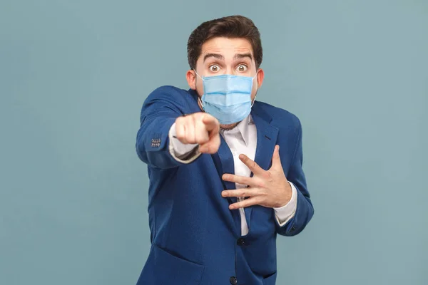 衝撃的な顔をしたカメラで外科用マスクで指を指差す衝撃的な男 ビジネスの人々の医学及びヘルスケアの概念 スタジオは青の背景で撮影 — ストック写真
