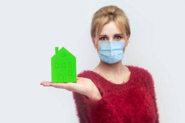 Evde kal, güvende kal. Karantina zamanı. Kırmızı bluzlu, ameliyat maskeli, elinde kağıt küçük bir ev tutan genç bir kadının portresi. Kapalı, izole, gri arka planda stüdyo çekimi