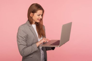Bilgisayarda yazı yazan akıllı ofis çalışanının portresi, takım elbiseli zarif pozitif iş kadını çalışmak için bilgisayar kullanıyor, internetten çalışıyor, uzaktan iş yapıyor. Stüdyo çekimi pembe arkaplanda izole edildi