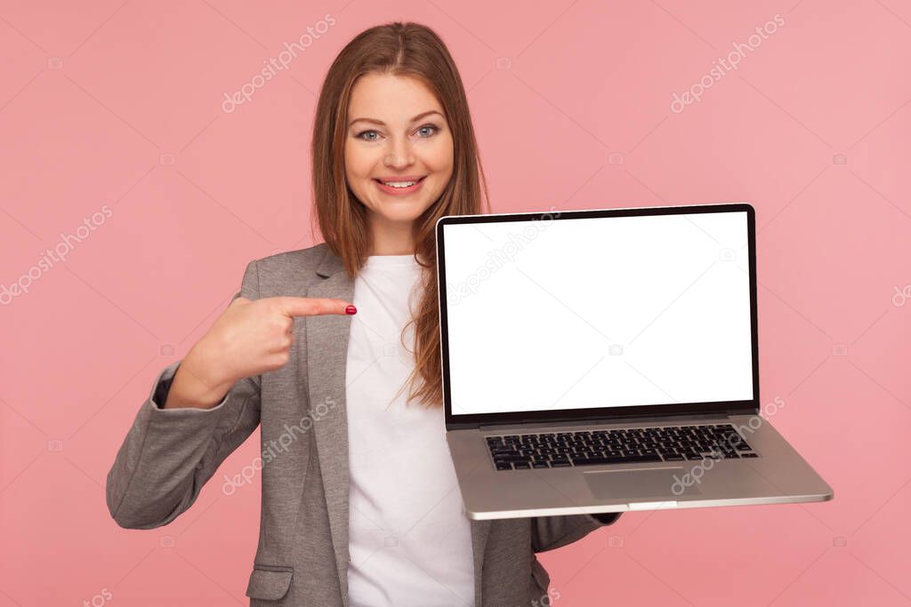 Portrait of beautiful elegant brunette businesswoman in suit jacket pointing at mock up display, holding laptop with blank screen for advertising. indoor studio shot isolated on pink background