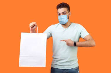 Neşeli alıcının portresi, tıbbi maskeli esmer adam gülümseyen ve paketleri işaret eden, alışveriş torbaları, tatil indirimlerine şaşıran. Turuncu arka planda kapalı stüdyo çekimleri