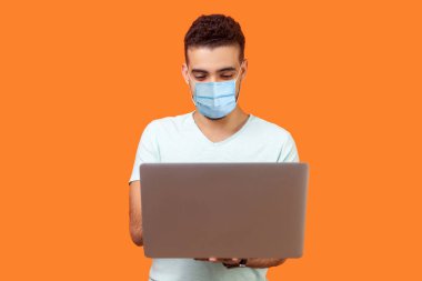 Cerrahi tıbbi maskeli neşeli esmer adamın portresi dizüstü bilgisayarda e-posta yazıyor ve gülümsüyor, olumlu mesaj okuyor ya da internette sörf yapıyor. Turuncu arka planda kapalı stüdyo çekimleri