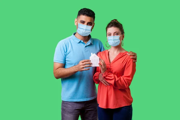 Stay home, stay safe. Portrait of safe young couple with surgical medical mask looking at camera with smile, both holding paper house and satisfied on quarantine time. isolated on green background