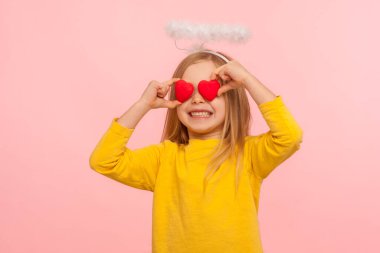 Portrait of nice angelic happy little girl with halo over head covering eyes with toy hearts and smiling sincerely, metaphor of look full of love. indoor studio shot isolated on pink background clipart