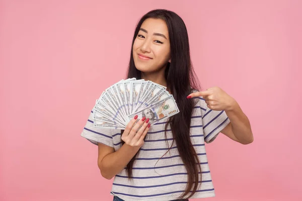 I\'m rich. Portrait of happy beautiful girl with brunette hair in striped t-shirt pointing at dollar bills, feeling wealthy successful, winning lottery. indoor studio shot isolated on pink background