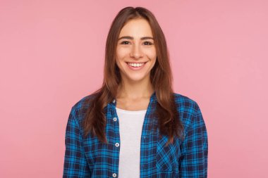Dost canlısı, kareli gömlekli, kameraya içtenlikle gülümseyen, hayattan memnun, pozitif haberler veren bir kızın portresi. Pembe arka planda kapalı stüdyo çekimleri