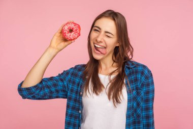 Şekerleme ve abur cubur. Ağzı açık, donut tutan ve tatlıyla eğlenen, kareli tişörtlü neşeli kızın portresi. Pembe arka planda kapalı stüdyo çekimleri